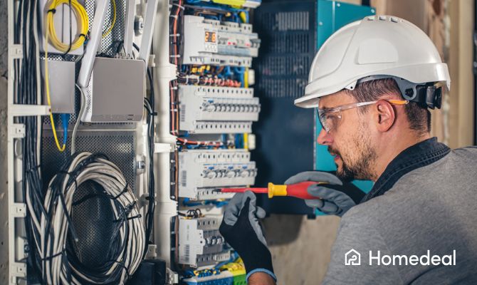 Elettricista al lavoro su un quadro elettrico, con casco e guanti protettivi, mentre installa componenti di un impianto elettrico domestico. - Homedeal