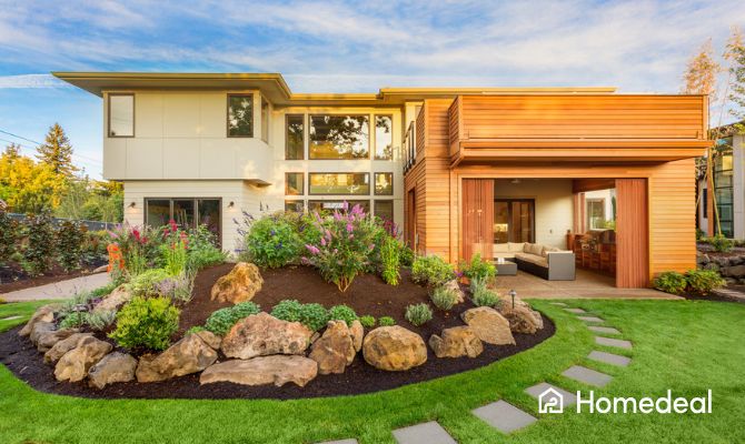 Giardino paesaggistico moderno con aiuola fiorita e vialetto in pietra, di fronte a una casa di design in legno e vetro.