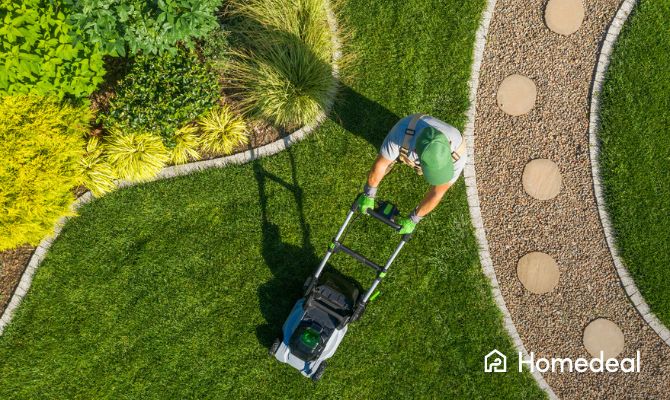 Vista aerea di un operatore che taglia l’erba in un giardino con vialetti curvilinei e piante decorative.