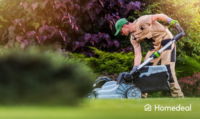 Operatore con tosaerba elettrico in fase di taglio dell’erba lungo bordure fiorite in un giardino residenziale.