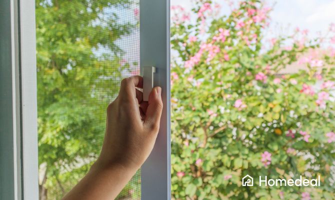 Mano che chiude una zanzariera su misura per finestra, con vista su giardino fiorito estivo - Homedeal
