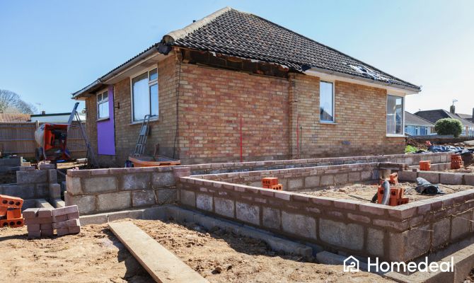 Ampliamento casa in corso con fondamenta in muratura e mattoni, accanto a un'abitazione esistente con tetto a spiovente; visibili attrezzi da cantiere, scale e betoniera sullo sfondo - Homedeal