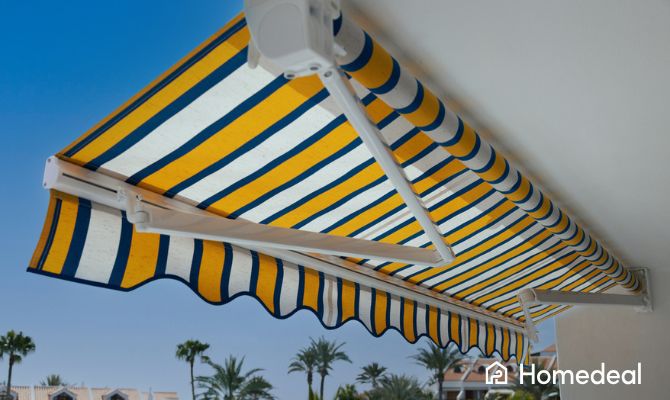Tenda da sole a bracci estensibili con telo a righe gialle e blu installata su terrazzo, con sfondo di palme e cielo azzurro - Homedeal