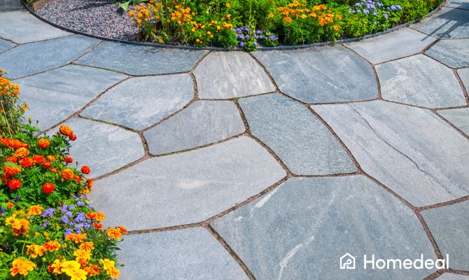 Vialetto da giardino con piastrelle per esterni in pietra naturale posate in modo irregolare, circondato da aiuole fiorite con margherite gialle, calendule arancioni e fiori viola - Homedeal